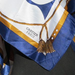 May include: A blue and gold silk scarf with a rope design and tassels. The scarf has the text 'CARTIER PARIS' printed on it.