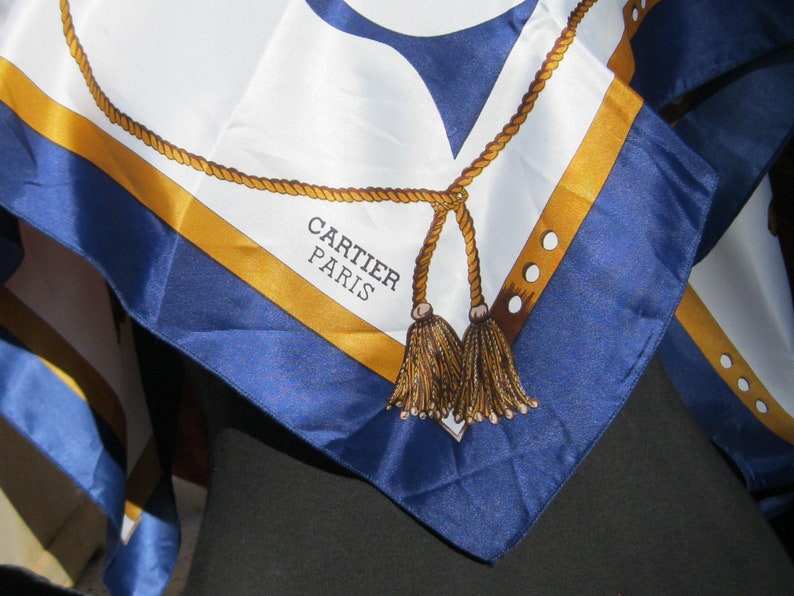 May include: A blue and gold silk scarf with a rope design and tassels. The scarf has the text 'CARTIER PARIS' printed on it.