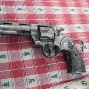 May include: A silver-colored revolver-shaped belt buckle with a black handle. The buckle is detailed with a cylinder, trigger, and hammer. The buckle is on a red and white checkered tablecloth.