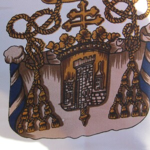 May include: A detailed illustration of a coat of arms with a gold rope border, a gold cross, a gold castle, and gold tassels. The castle is on a tan background with a blue and white striped border.