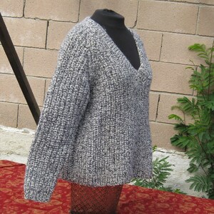 May include: A gray and white knit sweater with a deep V-neck. The sweater is displayed on a black mannequin and has a ribbed texture. The sleeves are long, and the overall style is casual and comfortable. The background includes a brick wall and greenery.