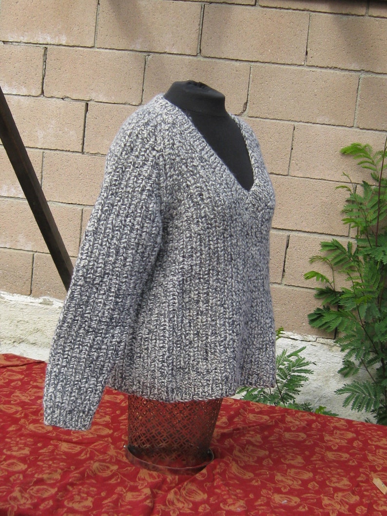 May include: A gray and white knit sweater with a deep V-neck. The sweater is displayed on a black mannequin and has a ribbed texture. The sleeves are long, and the overall style is casual and comfortable. The background includes a brick wall and greenery.