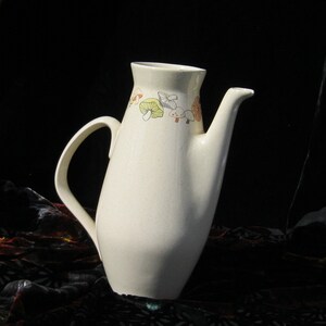 May include: Cream-colored ceramic pitcher with a long spout and curved handle. The pitcher is decorated with a band of colorful mushroom illustrations around the rim. The background is dark, highlighting the pitcher's shape and design.