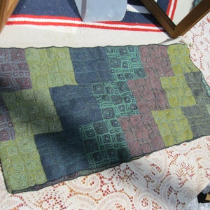 May include: A rectangular textile with a patchwork design. The color palette includes shades of green, blue, and purple. Each square features a repeating geometric pattern. The textile is likely a scarf or a small decorative piece.