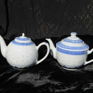 May include: Two white porcelain teapots with blue decorative bands and lids. The teapots have curved spouts and handles. One teapot has a round body, and the other has a more flattened shape. The teapots are on a black surface.