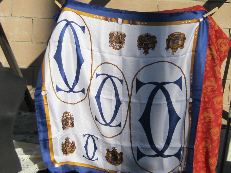 May include: A white silk scarf with a blue and gold design. The scarf features the Cartier logo in a blue and gold design. The scarf is folded and displayed on a brick wall background.