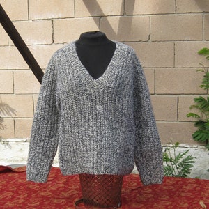 May include: A gray and white knit sweater with a V-neck design. The sweater is displayed on a black mannequin and is made of a thick, textured yarn. The sweater is displayed on a red floral patterned tablecloth.