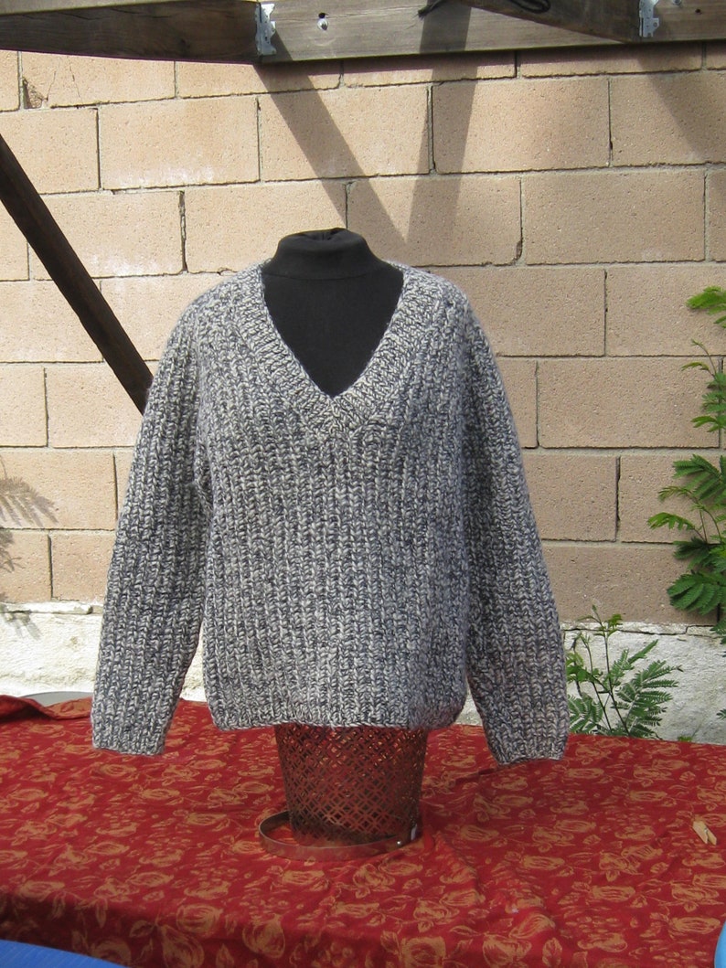 May include: A gray and white knit sweater with a V-neck design. The sweater is displayed on a black mannequin and is made of a thick, textured yarn. The sweater is displayed on a red floral patterned tablecloth.