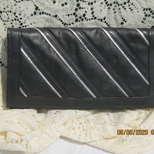 May include: Black clutch with diagonal silver stripes. The rectangular clutch is made of a smooth, dark material and features a flap closure. The silver stripes add a decorative element to the design. The clutch is a stylish accessory.