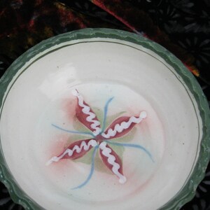 May include: A round, off-white ceramic bowl with a scalloped, translucent green rim. The interior features a hand-painted, five-petaled floral design in shades of muted red, soft green, and blue, accented with a white, wavy, raised-texture line.