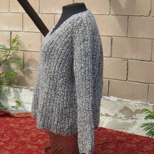 May include: A gray and white knit sweater with a V-neck design. The sweater is displayed on a black mannequin and has a ribbed texture. The garment is made of a thick yarn and appears warm and cozy.