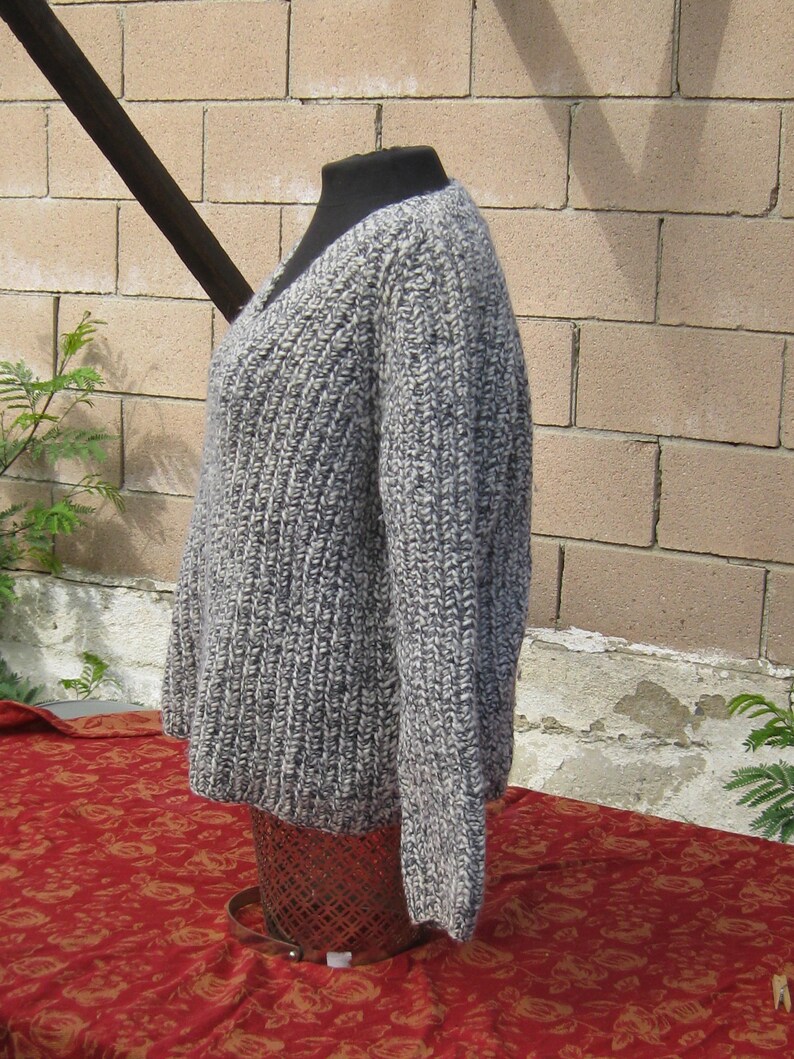 May include: A gray and white knit sweater with a V-neck design. The sweater is displayed on a black mannequin and has a ribbed texture. The garment is made of a thick yarn and appears warm and cozy.