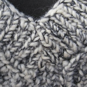 May include: Close-up of a hand-knitted textile with a chunky, textured appearance. The yarn is a mix of white and dark gray, creating a speckled effect. The stitches are large and visible, showing the handmade quality of the item.