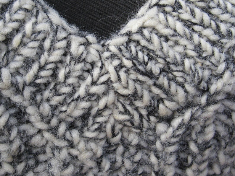 May include: Close-up of a hand-knitted textile with a chunky, textured appearance. The yarn is a mix of white and dark gray, creating a speckled effect. The stitches are large and visible, showing the handmade quality of the item.