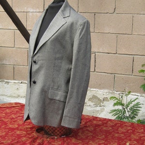 May include: A gray and white houndstooth blazer with a notched lapel and two black buttons. The blazer is displayed on a black mannequin and set against a brick wall. The blazer is on a red floral tablecloth.