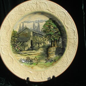 May include: A decorative ceramic plate with a detailed landscape scene. The plate features a cream-colored border with embossed floral patterns. The central image depicts a building with trees and a stone bridge, painted in muted tones.