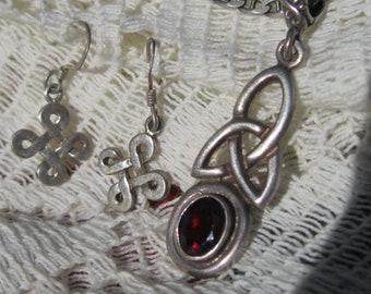 May include: A silver jewelry set featuring Celtic knot earrings and a pendant with a red gemstone. The earrings have a looped design, and the pendant has an oval-shaped stone. The jewelry is displayed on a white, textured surface.