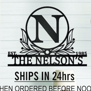 May include: Black metal family name sign with the letter "N" in a circle, "EST. 1985" and "THE NELSON'S" below. The sign has a decorative leaf design. The sign is mounted on a white wooden wall.