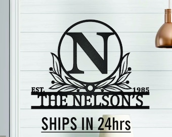 Puede incluir: Un letrero de monograma de metal negro mate con la letra N en un círculo, rodeado de ramas frondosas. El letrero dice "EST. THE NELSON'S 1985".