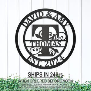 May include: Black metal round sign with the names David & Amy, the initial T for Thomas, and the year 2024. The sign has a decorative floral design.