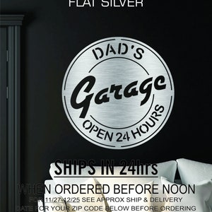 Pode incluir: Um sinal de metal prateado com o texto "DAD'S Garage OPEN 24 HOURS" em um c&iacute;rculo. O sinal est&aacute; montado em uma parede preta.