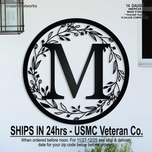 May include: Black metal wall art featuring the letter "M" inside a circle, surrounded by a floral wreath design. The text "14 GAUGE AMERICAN MADE STEEL" is visible. The product is from "crossmetalworks".