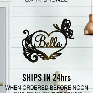 May include: A black metal wall decor with a heart shape and a butterfly design. The heart has the name "Bella" written inside. The text "SHIPS IN 24hrs" and "WHEN ORDERED BEFORE NOON" is also visible.
