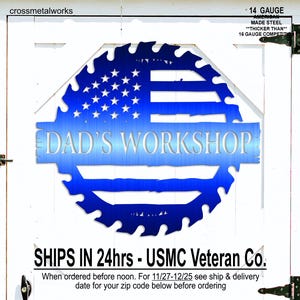 Op de afbeelding: Een blauw metalen bord met de tekst "DAD'S WORKSHOP" in een gestileerd lettertype. Het bord heeft de vorm van een zaagblad en heeft het ontwerp van de Amerikaanse vlag. Het bord is gemaakt van 14 gauge staal.