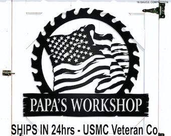 May include: Black metal sign featuring an American flag design within a saw blade outline. The sign reads "PAPA'S WORKSHOP" and includes text indicating it's made of American steel. The sign is mounted on a white door.
