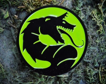 May include: A black and green enamel pin featuring a stylized image of a snarling creature with sharp teeth. The creature is depicted in a circular shape with a green background.