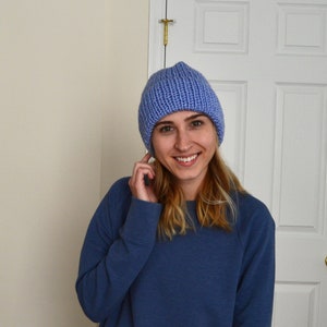 May include: A light blue knit beanie hat. The hat is made of a thick, ribbed knit material. The person in the image is wearing a blue sweatshirt. The background is a plain wall and a white door.