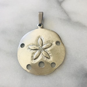 May include: A silver-toned pendant featuring a sand dollar design. The pendant has a circular shape with a raised, five-petal flower in the center and several small holes around the edge. The pendant has a loop at the top for a chain.