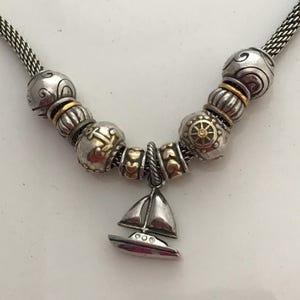 May include: A silver-toned chain necklace featuring a collection of beads with gold-toned accents. The beads include nautical motifs such as an anchor and a ship's wheel. A sailboat charm hangs from the centre, accented with small clear stones.