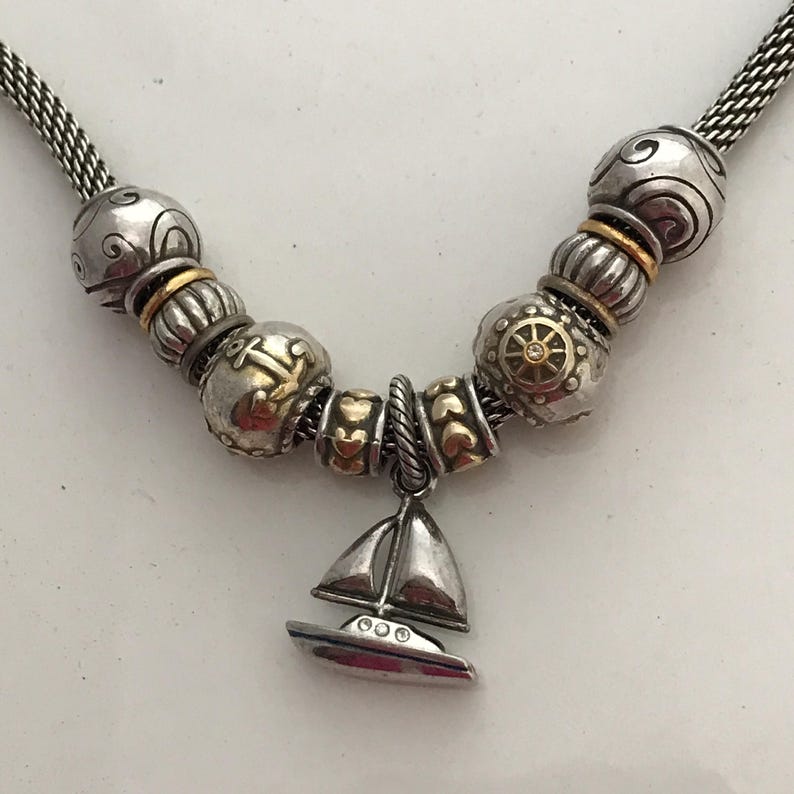 May include: A silver-toned chain necklace featuring a collection of beads with gold-toned accents. The beads include nautical motifs such as an anchor and a ship's wheel. A sailboat charm hangs from the centre, accented with small clear stones.