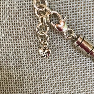 May include: A silver-toned metal necklace chain featuring a lobster claw clasp and a small heart-shaped charm dangling from the end. The charm is embossed with a stylized letter B. The chain consists of circular links connected to a cylindrical component.