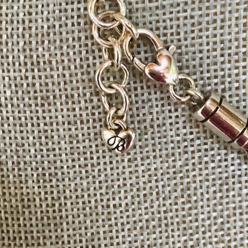 May include: A silver-toned metal necklace chain featuring a lobster claw clasp and a small heart-shaped charm dangling from the end. The charm is embossed with a stylized letter B. The chain consists of circular links connected to a cylindrical component.