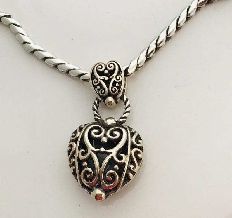 May include: A silver-toned necklace featuring a heart-shaped pendant with intricate scrollwork and cut-out details. The pendant hangs from a twisted silver-toned chain by a matching decorative bail, showcasing a polished metallic finish.