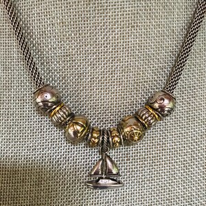 May include: A silver-toned chain necklace featuring a series of metallic beads with gold-toned accents and a central sailboat charm. The beads display various embossed patterns, including swirls and geometric shapes, suspended on a woven metal chain.