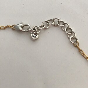 Puede incluir: Un cierre de collar con un mosquet&oacute;n plateado y una serie de eslabones circulares de ajuste. Un peque&ntilde;o dije en forma de coraz&oacute;n con las letras "JB" grabadas cuelga del cierre, conectado a un segmento de cadena de eslabones dorados.
