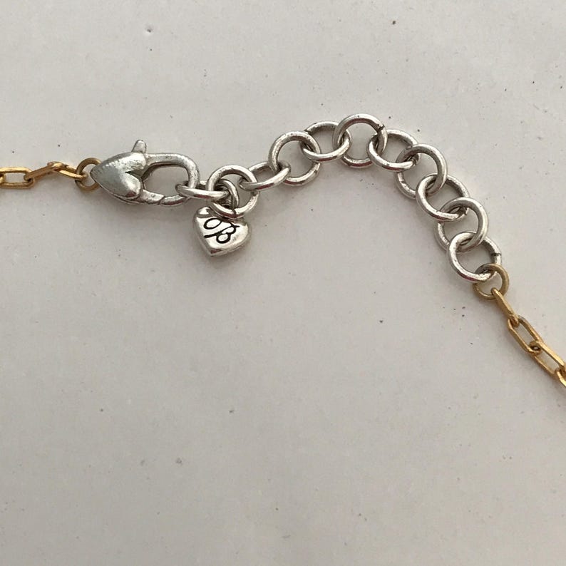 Puede incluir: Un cierre de collar con un mosquet&oacute;n plateado y una serie de eslabones circulares de ajuste. Un peque&ntilde;o dije en forma de coraz&oacute;n con las letras "JB" grabadas cuelga del cierre, conectado a un segmento de cadena de eslabones dorados.