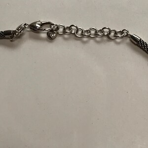 May include: A silver-toned necklace featuring a woven mesh chain, a lobster claw clasp, and an adjustable extender chain with circular links. A small heart-shaped charm with the letters GB is attached near the clasp mechanism of the jewellery.