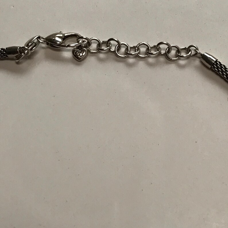 May include: A silver-toned necklace featuring a woven mesh chain, a lobster claw clasp, and an adjustable extender chain with circular links. A small heart-shaped charm with the letters GB is attached near the clasp mechanism of the jewellery.