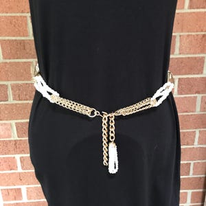 May include: Gold-tone chain belt with white beaded accents and a dangling tassel. The belt is worn over a black dress, creating a contrast with the brick background. The belt is a stylish accessory.