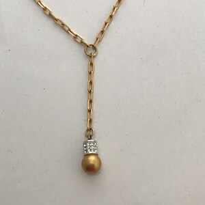 Puede incluir: Un collar de tono dorado con un dise&ntilde;o de cadena en forma de Y con eslabones rectangulares alargados. El colgante central termina en una esfera con acabado dorado mate, acentuada por una banda plateada incrustada con peque&ntilde;os cristales.