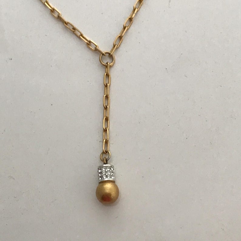 Puede incluir: Un collar de tono dorado con un dise&ntilde;o de cadena en forma de Y con eslabones rectangulares alargados. El colgante central termina en una esfera con acabado dorado mate, acentuada por una banda plateada incrustada con peque&ntilde;os cristales.