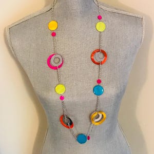 May include: A long necklace with a silver chain featuring colorful circular and round pendants. The pendants are in shades of yellow, pink, orange, blue, and gray. The necklace is displayed on a mannequin.