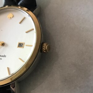 May include: A close-up of a Kate Spade watch with a white face, gold-tone bezel, and a black leather strap. The watch has gold-tone hour markers, a date window, and a gold spade logo. The crown is gold-tone.