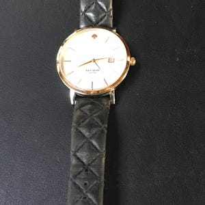 May include: A gold-tone Kate Spade watch with a white face, gold hands, and a date window. The watch has a black quilted leather strap. The watch is displayed on a black surface.