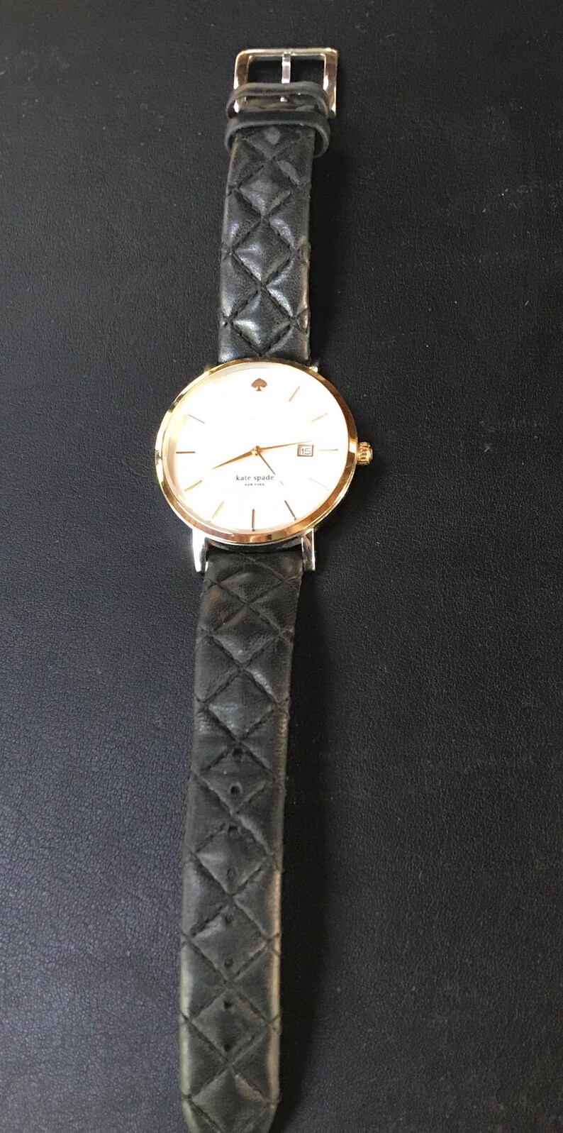 May include: A gold-tone Kate Spade watch with a white face, gold hands, and a date window. The watch has a black quilted leather strap. The watch is displayed on a black surface.