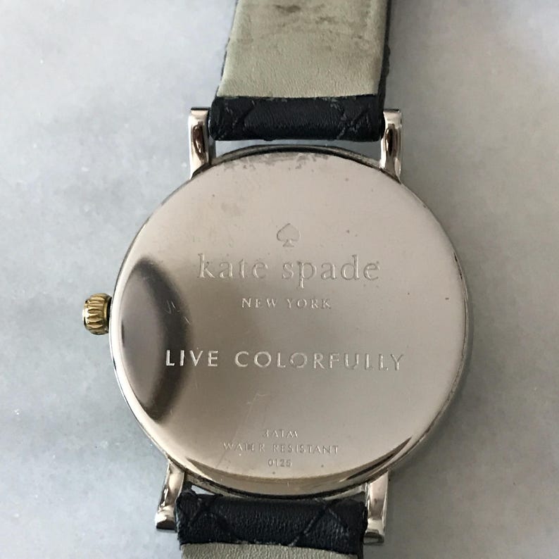May include: A silver Kate Spade watch with a black leather strap. The watch face is round and engraved with "kate spade NEW YORK" and "LIVE COLORFULLY". The watch has a gold-coloured crown.